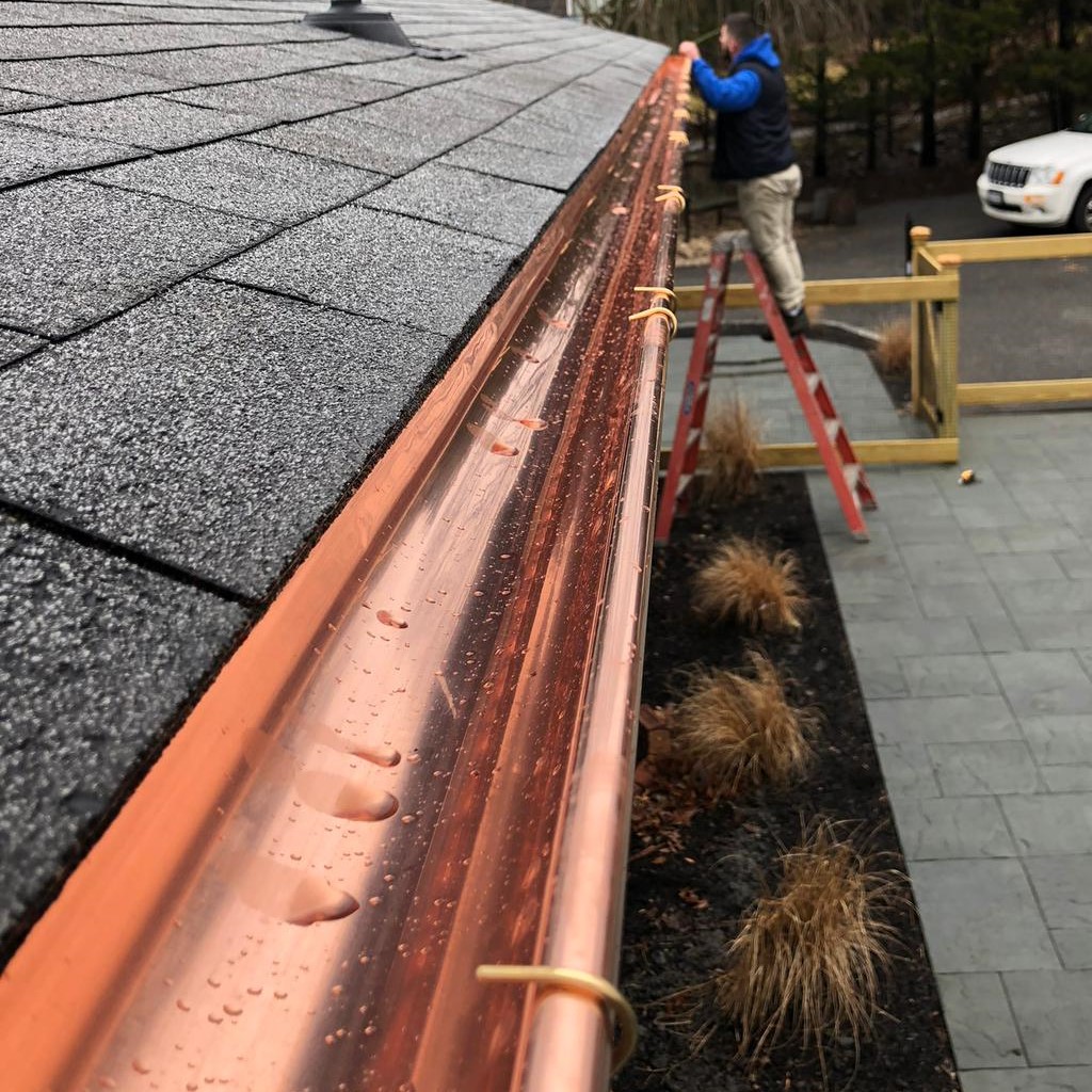Copper gutter installation on a residential roof by a professional roofer standing on a ladder.